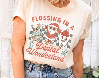 Könnte beinhalten: Cremefarbenes T-Shirt mit dem Schriftzug "FLOSSING IN A Dental Wonderland" in roter Schrift. Die Grafik zeigt einen Weihnachtsbaum, den Weihnachtsmann und einen Zahn, alle mit festlichem Zubehör. Das Shirt ist aus weichem Material.