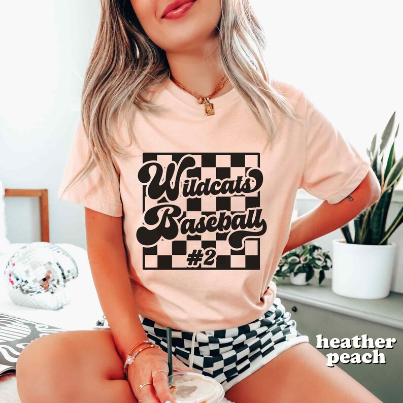 May include: Heather peach t-shirt with a black and white checkered design. The shirt features the words "Wildcats Baseball #2" in a retro font. The model is wearing a gold necklace and bracelets.
