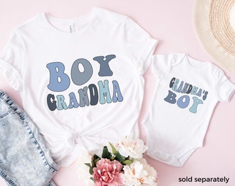 May include: Two white t-shirts with blue and gray text. The larger shirt says "Boy Grandma" and the smaller shirt says "Grandma's Boy".