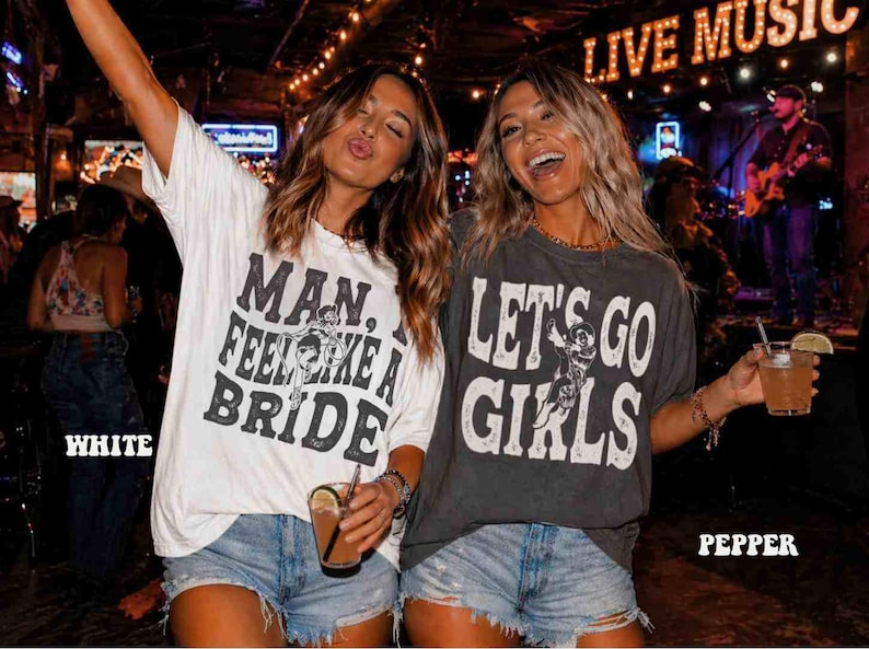 K&ouml;nnte beinhalten: Zwei Frauen in einem Bar-Ambiente, tragen Grafik-T-Shirts und Jeansshorts, l&auml;cheln und halten Getr&auml;nke. Ein Shirt tr&auml;gt die Aufschrift "MAN. FEEL LIKE A BRIDE", das andere "LET'S GO GIRLS". Im Hintergrund ein Schild mit "LIVE MUSIC".