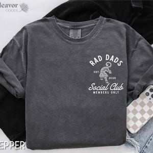 May include: A dark gray t-shirt with a white graphic design that says "Rad Dads Social Club Members Only" with a tiger illustration and the text "Est. 2025".