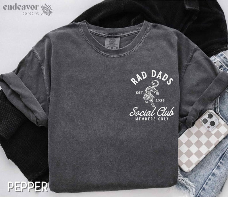May include: A dark gray t-shirt with a white graphic design that says "Rad Dads Social Club Members Only" with a tiger illustration and the text "Est. 2025".