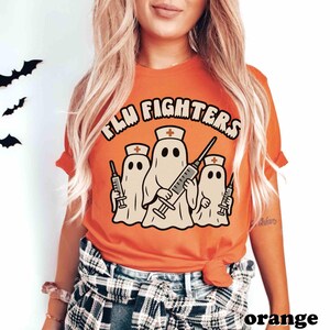 May include: An orange t-shirt featuring the words "FLU FIGHTERS" above a graphic of three ghost figures dressed as nurses, each holding a syringe. The tee is a solid orange colour.