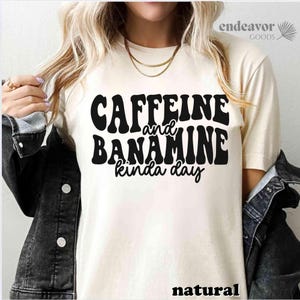May include: A cream-coloured t-shirt featuring the text "Caffeine and Banamine kinda day" in bold, black, retro lettering. The word "natural" is printed at the bottom. The shirt is from the Endeavor Goods collection.