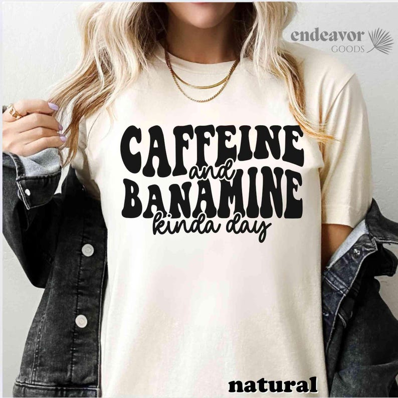 May include: A cream-coloured t-shirt featuring the text "Caffeine and Banamine kinda day" in bold, black, retro lettering. The word "natural" is printed at the bottom. The shirt is from the Endeavor Goods collection.