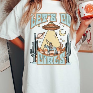 以下が含まれることがあります： UFO、馬に乗ったカウガール、そして「Let's Go Girls」というテキストが描かれた白いTシャツです。