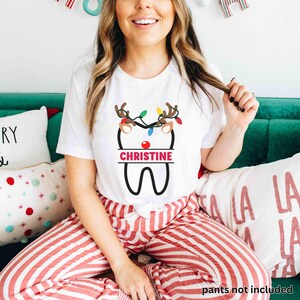 以下が含まれることがあります： クリスマスをテーマにした歯のデザインが施された白いTシャツ。デザインには、トナカイの角、カラフルなライト、ピンクのバナーに「CHRISTINE」という名前が含まれています。 赤と白のストライプのパジャマパンツを着用しています。
