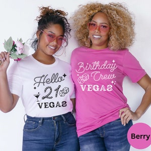 May include: Two women wearing matching t-shirts. The woman on the left is wearing a white t-shirt with the text "Hello 21 Vegas" in black. The woman on the right is wearing a pink t-shirt with the text "Birthday Crew Vegas" in white.
