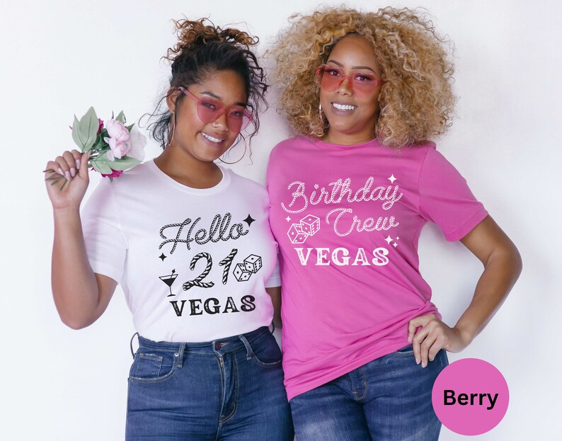 May include: Two women wearing matching t-shirts. The woman on the left is wearing a white t-shirt with the text "Hello 21 Vegas" in black. The woman on the right is wearing a pink t-shirt with the text "Birthday Crew Vegas" in white.