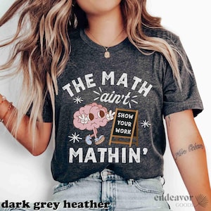 May include: A dark grey heather t-shirt with a white graphic of a brain holding a sign that says "Show Your Work" and the text "The Math ain't Mathin'"