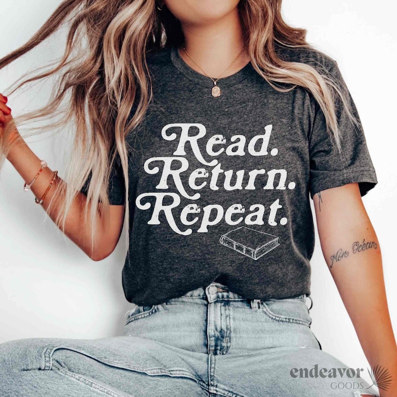 May include: Dark gray t-shirt with the text "Read. Return. Repeat." in a white, stylized font. A small book illustration is below the text. The shirt is a crew neck and appears to be made of a soft material.