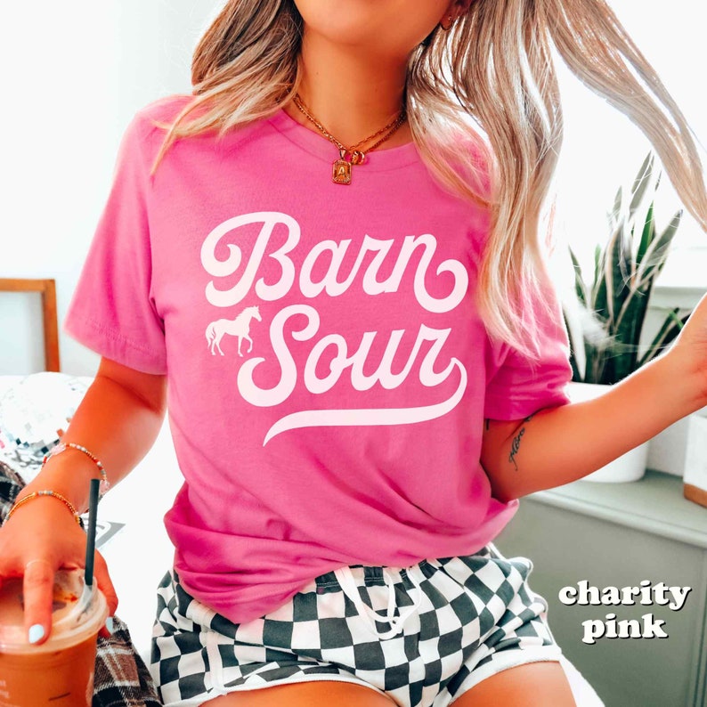 May include: A pink t-shirt with the text "Barn Sour" in white cursive lettering. A small white silhouette of a horse is to the left of the text. The t-shirt is being worn by a person wearing black and white checkered shorts.