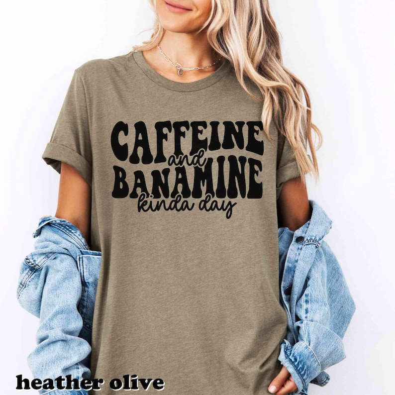 May include: Heather olive green t-shirt with the text "CAFFEINE and BANAMINE kinda day" in black. The shirt has short sleeves and a crew neck. The words "heather olive" are at the bottom of the image.