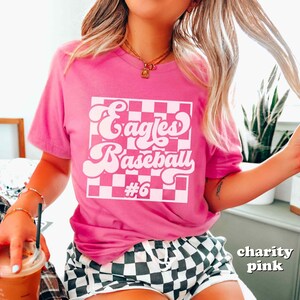 May include: A pink t-shirt with the words "Eagles Baseball #6" in a white retro font and a checkered background. The shirt is a solid pink color.