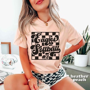 May include: A pink t-shirt with a black and white checkered background and the text "Eagles Softball #2" in a retro font.