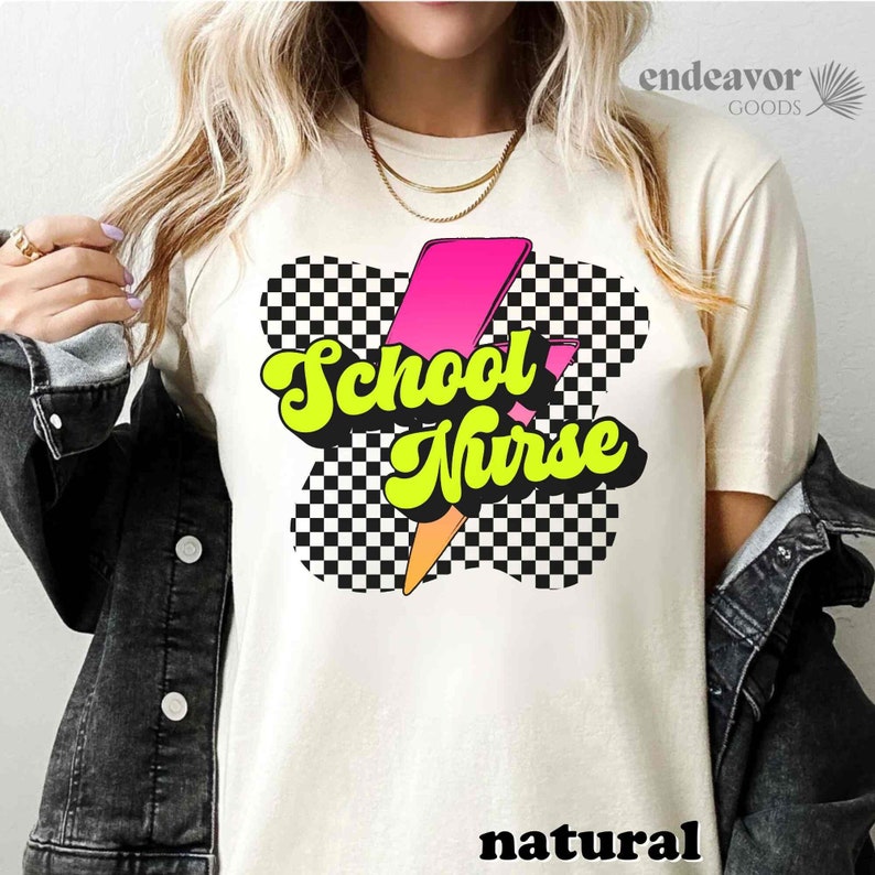 Op de afbeelding: Een wit T-shirt met een zwart-wit geruit ontwerp en een roze en gele bliksemgrafiek. De tekst "School Nurse" is geschreven in een retro-lettertype.