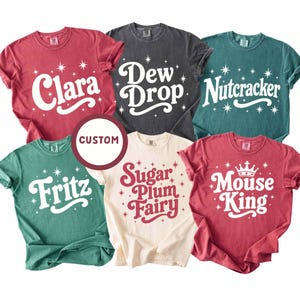 May include: Six short-sleeved crewneck t-shirts in red, charcoal, teal, and cream colors. Each shirt features white or red text including Clara, Dew Drop, Nutcracker, Fritz, Sugar Plum Fairy, and Mouse King, accented with star graphics and a custom circle.