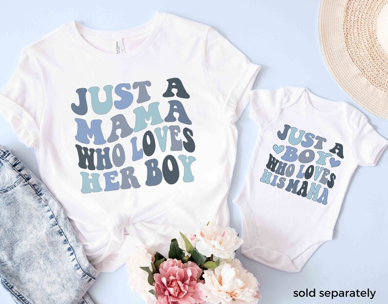 May include: A white t-shirt with the text "Just a Mama who loves her boy" in blue and gray letters. A white baby onesie with the text "Just a Boy who loves his Mama" in blue and gray letters. Both shirts have a heart design.