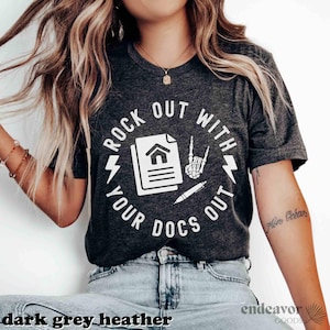 May include: Dark grey heather t-shirt with a white graphic. The graphic features the text "ROCK OUT WITH YOUR DOCS OUT" in a circular design, along with a house document, a pen, and a skeleton hand making a rock and roll gesture.