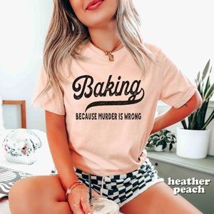 Pode incluir: Uma camiseta rosa com um gr&aacute;fico preto de estilo vintage que diz "Baking because murder is wrong".