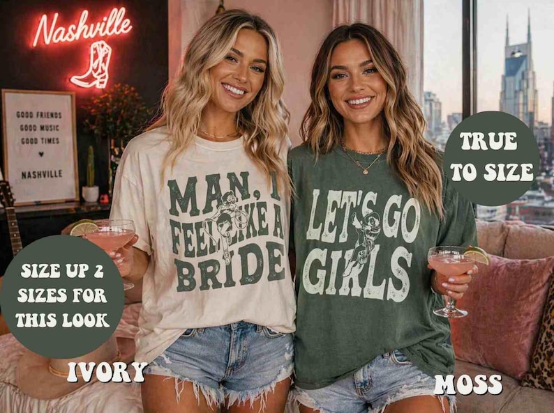 K&ouml;nnte beinhalten: Zwei Frauen tragen Grafik-T-Shirts und Jeansshorts. Eines ist elfenbeinfarben mit dem Text "MAN, I FEEL LIKE A BRIDE", das andere ist moosgr&uuml;n mit "LET'S GO GIRLS". Im Hintergrund ein Neon-Schild "Nashville" und ein gerahmtes Schild.