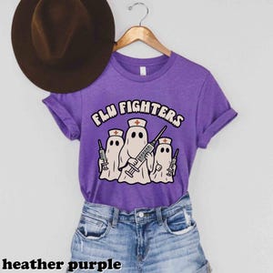 May include: Heather purple t-shirt featuring the text "FLU FIGHTERS" and a graphic of three ghost figures wearing nurse hats and holding syringes. The t-shirt is displayed on a wooden hanger.