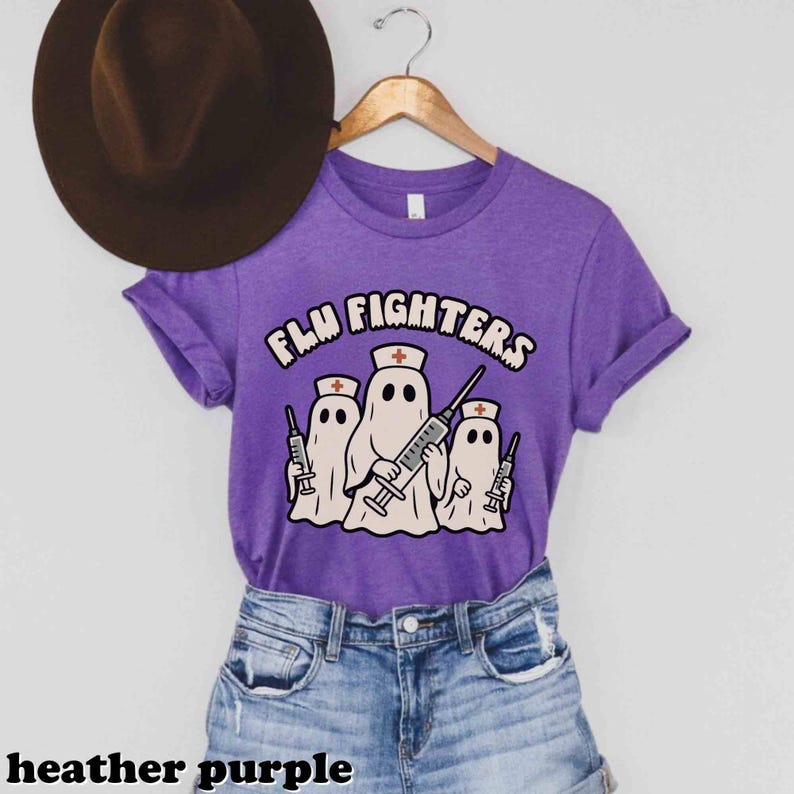 May include: Heather purple t-shirt featuring the text "FLU FIGHTERS" and a graphic of three ghost figures wearing nurse hats and holding syringes. The t-shirt is displayed on a wooden hanger.