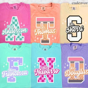 May include: A collection of six colorful t-shirts, each with a unique design featuring a large letter and a name. The shirts are in shades of pink, orange, yellow, green, blue, and purple. The designs include the names "Anderson", "Thomas", "Sherri", "Francesca", "Navarro", and "Douglas".