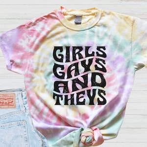 May include: A tie-dye t-shirt with the text "Girls, Gays, and Theys" in black lettering. The shirt is a light yellow, pink, and blue color.