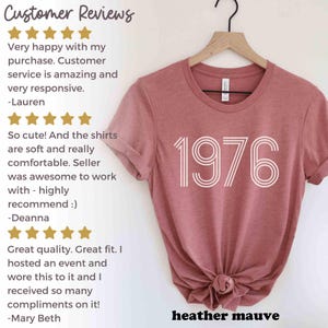 May include: Mauve heather t-shirt featuring the year "1976" in white. The tee is hanging on a wooden hanger. The words "heather mauve" are printed at the bottom of the shirt.