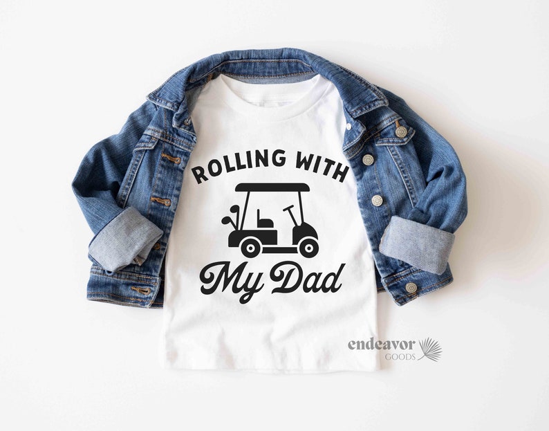 May include: White t-shirt with black text that reads "Rolling with My Dad" and a black silhouette of a golf cart.