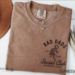May include: A brown t-shirt with a vintage-style graphic design that says "Rad Dads Social Club Members Only" with a tiger illustration and the text "Est. 2025".