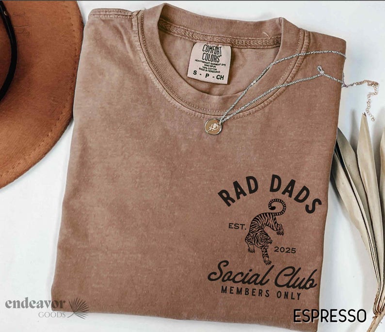 May include: A brown t-shirt with a vintage-style graphic design that says "Rad Dads Social Club Members Only" with a tiger illustration and the text "Est. 2025".