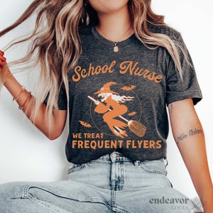 May include: A dark gray t-shirt with the text "School Nurse" in orange lettering. A witch flying on a broomstick is depicted in an orange circle with the text "We Treat Frequent Flyers" below. The text "Mrs. Ocean" is written in cursive on the right side of the shirt.
