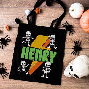 May include: Black tote bag with a Halloween theme. The bag features a green and orange lightning bolt design with the name "HENRY" in green. Four cartoon skeletons are dancing around the lightning bolt. Decorative spiders, pumpkins, and a skull are also present.