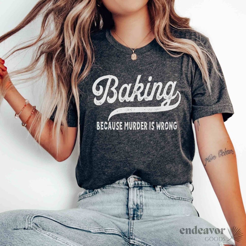 Pode incluir: Uma camiseta cinza escura com um gr&aacute;fico branco de estilo vintage que diz "Baking because murder is wrong".