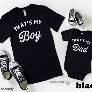 May include: Black t-shirts with white text. The larger shirt says "That's My Boy" and the smaller shirt says "That's My Dad".