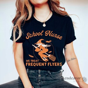 May include: A black t-shirt with an orange graphic of a witch flying on a broomstick. The text "School Nurse" is above the witch and "We Treat Frequent Flyers" is below the witch.