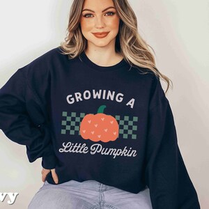 May include: Navy blue sweatshirt with the text "GROWING A Little Pumpkin" and a pumpkin graphic. The pumpkin is orange with green accents and is centered on a checkered background. The word "navy" is printed at the bottom left.