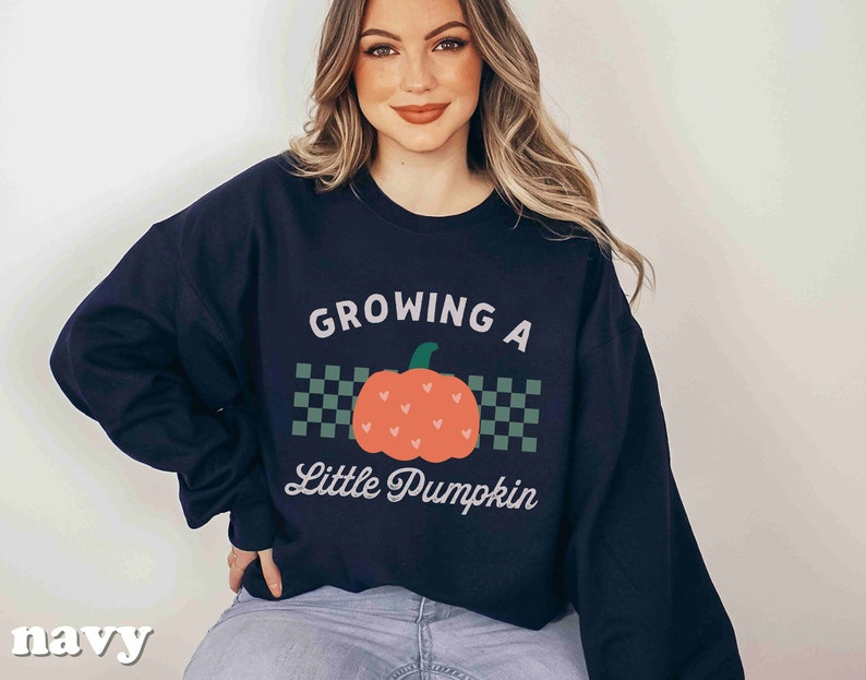 May include: Navy blue sweatshirt with the text "GROWING A Little Pumpkin" and a pumpkin graphic. The pumpkin is orange with green accents and is centered on a checkered background. The word "navy" is printed at the bottom left.