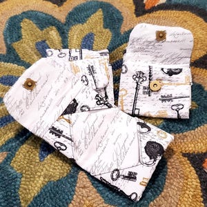 May include: White fabric wallets with a vintage key and script design. The wallets have a gold snap closure. The design includes black and gold keys and script on a white background. The wallets are open and closed.