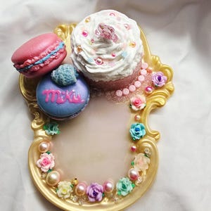 Op de afbeelding: Een goudkleurig decoratief dienblad versierd met kleine kleurrijke roosjes en parels, met een roze macaron, een paarse macaron met de tekst Miku, en een witte cupcake met strooisel en een klein figuurtje, met de tekst HATSUNE erop.