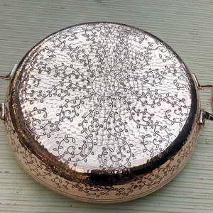May include: A copper-colored, round cooking pan with two handles. The pan features a hammered texture and an intricate floral design etched into the bottom. The handles are curved and attached to the sides of the pan.