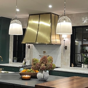 May include: A modern kitchen with a gold metal range hood, white pendant lights, and a dark green island with a farmhouse sink. The kitchen features a marble backsplash and a window overlooking a backyard.