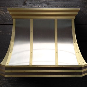 May include: A stainless steel range hood with a curved top and brass trim. The hood has three vertical brass strips with decorative rivets. The bottom edge of the hood has two bands of brass trim with decorative rivets.