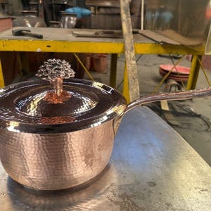 May include: A copper saucepan with a lid and a long handle. The pot has a hammered texture and a decorative knob on the lid. The handle is curved and attached to the side of the pot. The pot is sitting on a metal surface.