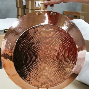 May include: A large, hammered copper tray with two handles. The tray has a circular shape with a textured surface. The handles are made of the same copper material. The tray is being held by a person.