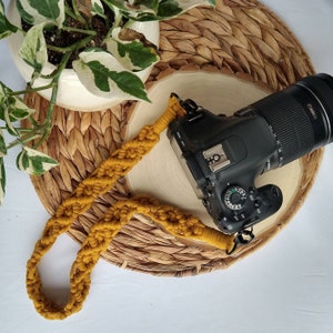 May include: A mustard yellow braided camera strap with a black camera attached. The camera has a black lens with the word "Canon" printed on it.