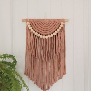 May include: A blush pink macrame wall hanging with wooden beads. The hanging features a half-circle design with fringe at the bottom.