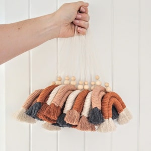 May include: A set of 10 colorful macrame wall hangings with wooden beads. Each hanging features a rainbow of colors, including orange, brown, cream, and black. The hangings are tied together with a white string.